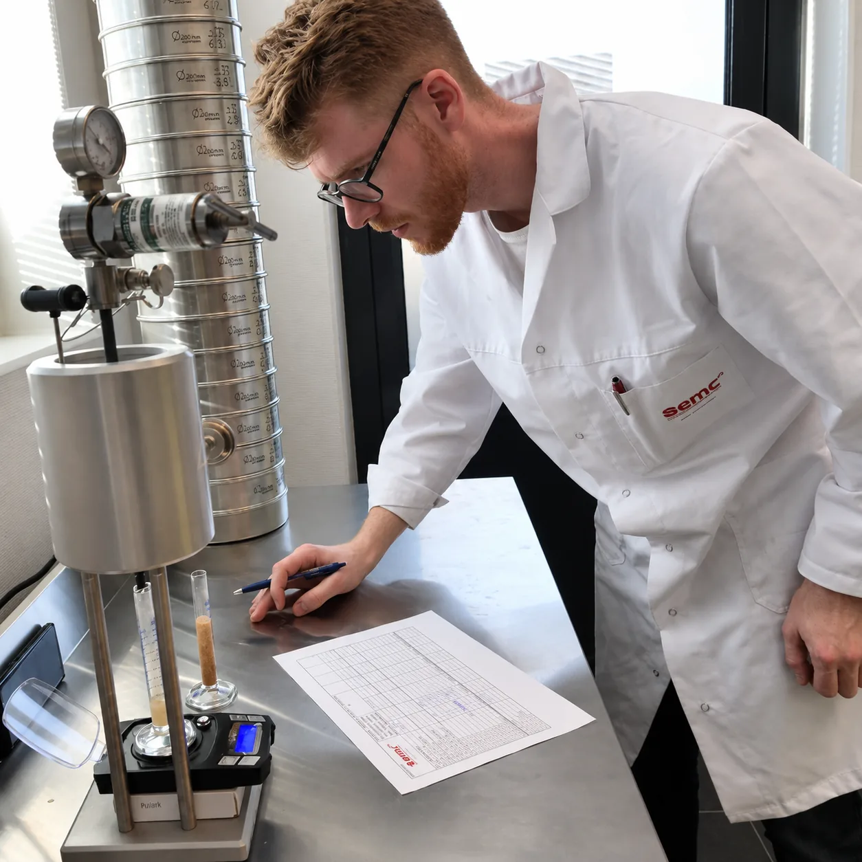 Laboratorietekniker noterer resultater ved siden av en stabel med testsikter